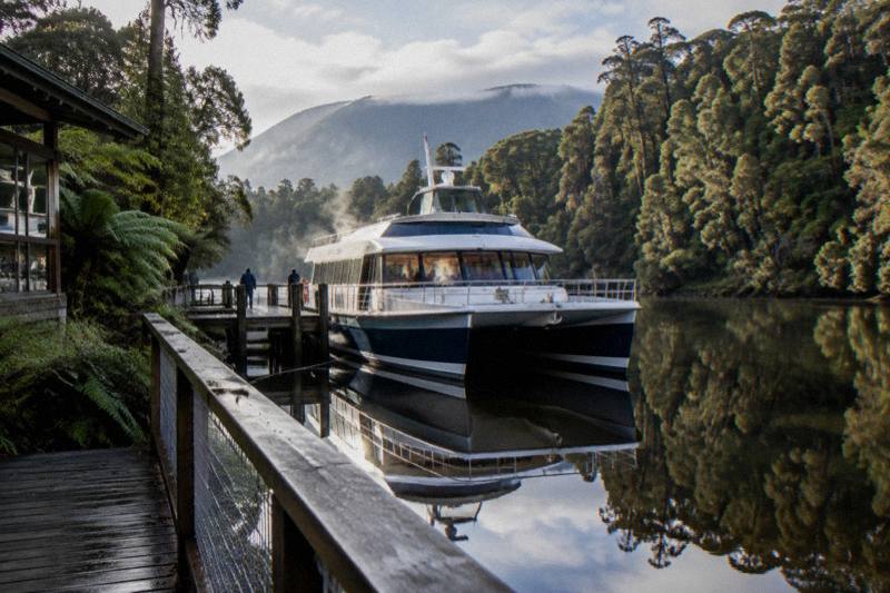 A passenger catamaran cruising the Gordon River in Tasmania on a morning lunch cruise, with mirror-still water and rainforest on both banks