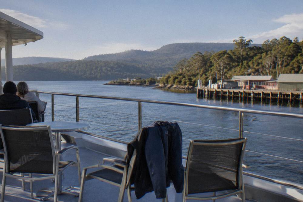 A cruise boat upper deck with outdoor seating and a rail overlooking a wide harbour
