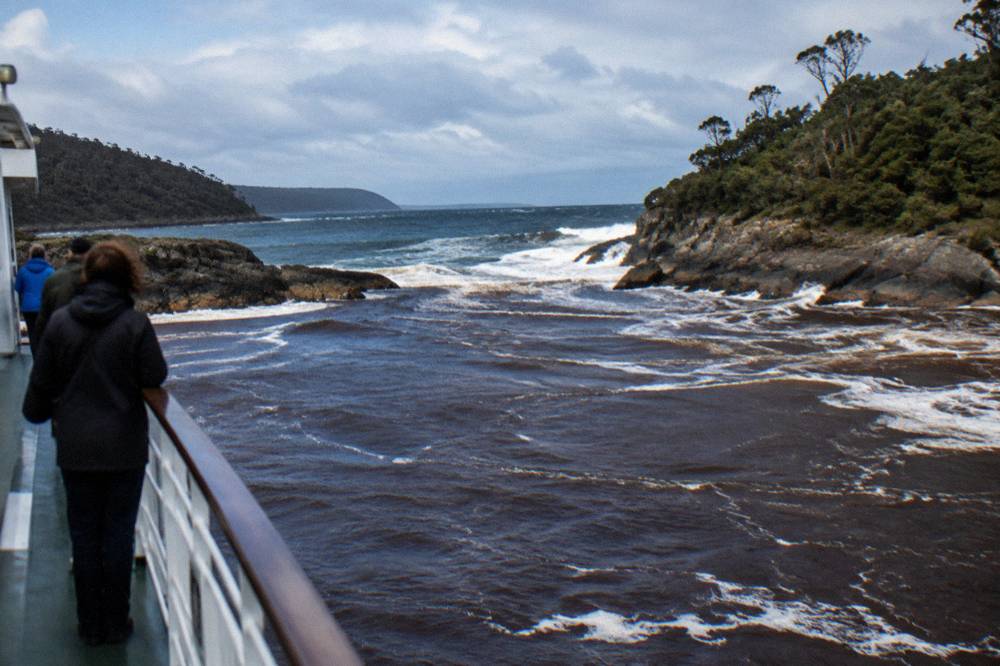 Dark tannin-stained harbour water meeting ocean swell at a narrow rocky channel entrance