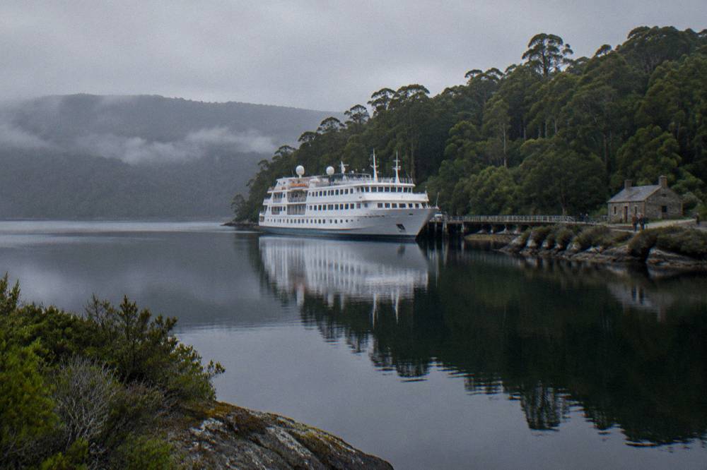 A cruise vessel moored beside a green forested island under a grey sky on dark still water