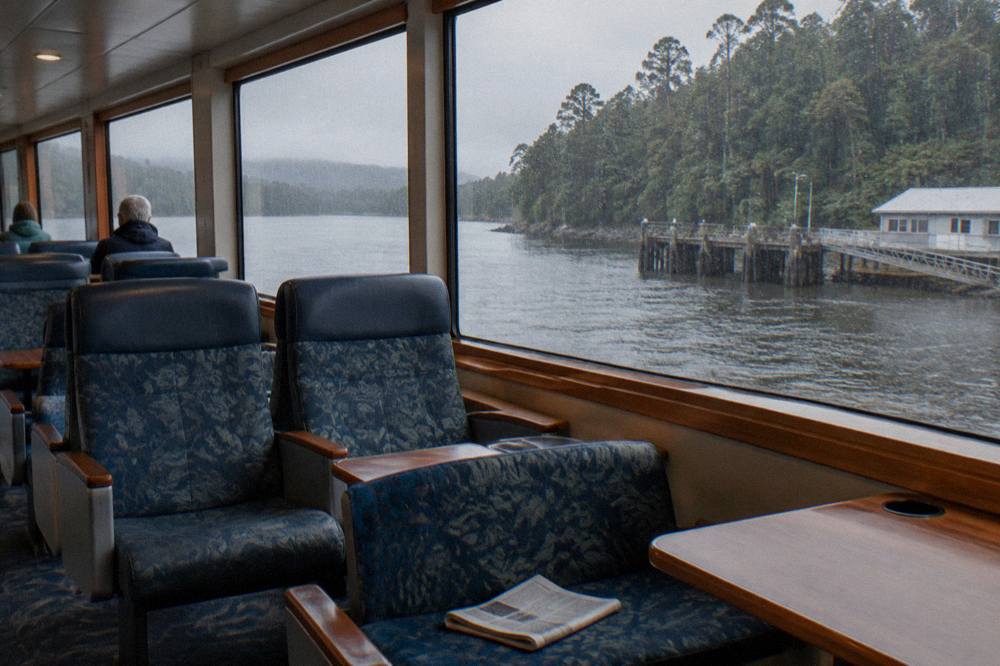 Timber cruise boat deck with rows of seats beside large windows overlooking a rainforest-lined river.