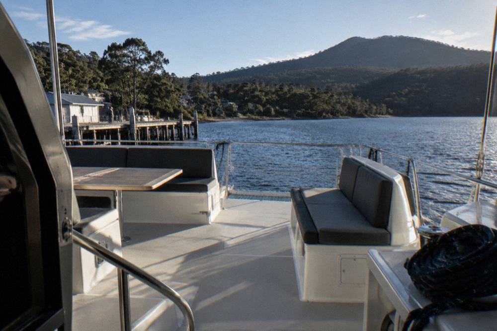 Upper deck of a catamaran with outdoor seating and a view across dark harbour water toward forested hills.