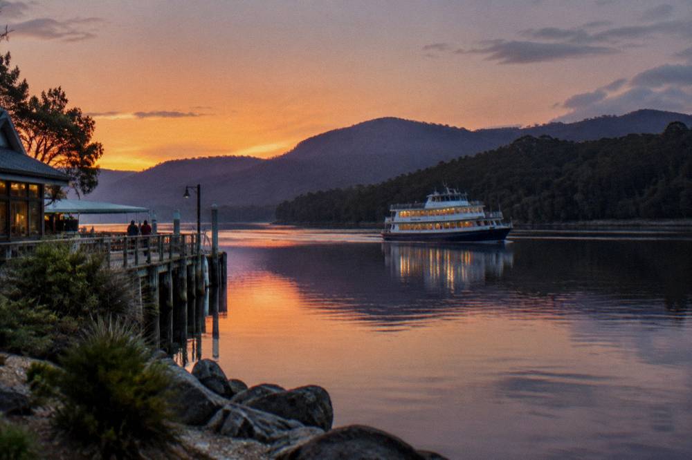 A cruise boat on calm dark water in late afternoon light with forested hills behind it.